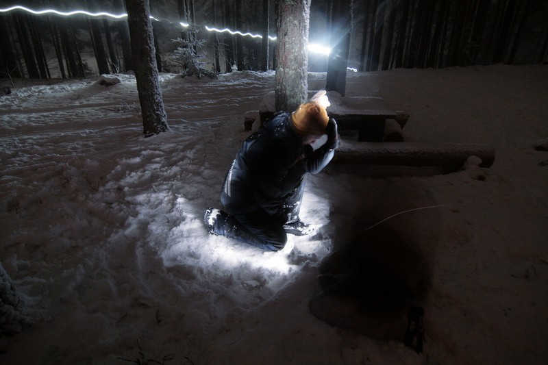 Podświetlona klęcząca postać na śniegu w lesie.